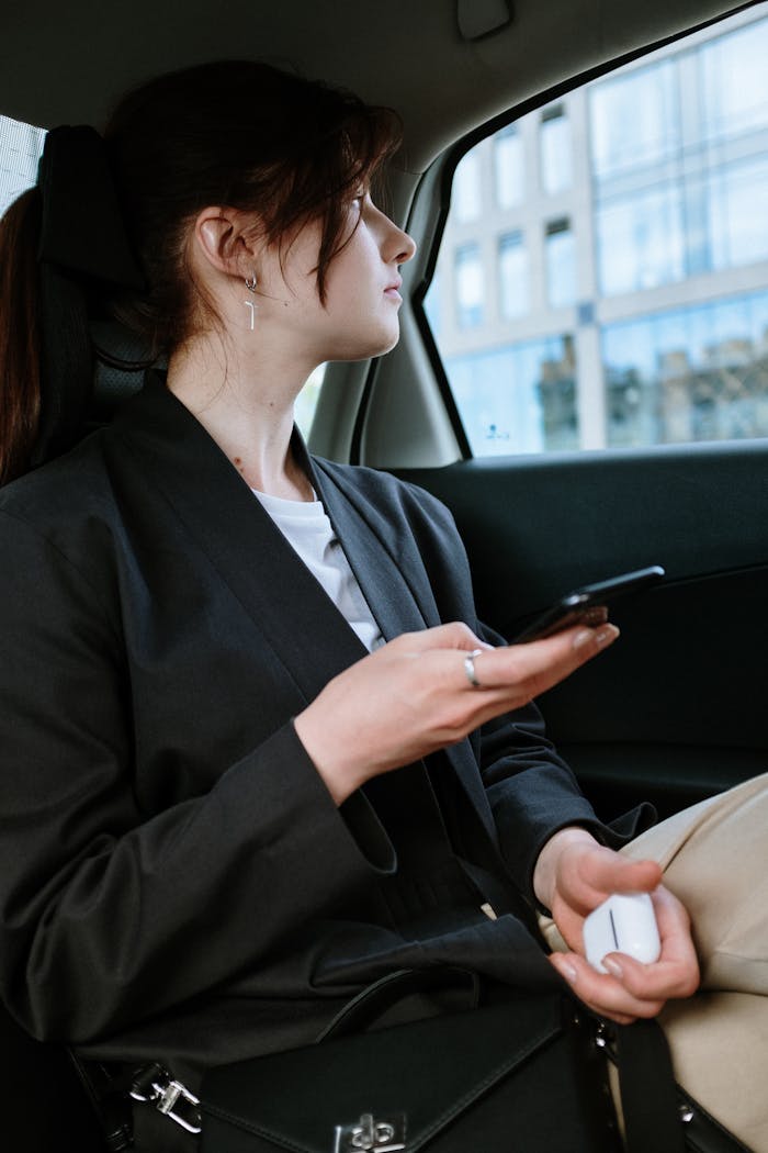 our-services-2 A woman with a ponytail and earrings uses a smartphone in the back of a car, focusing outside.